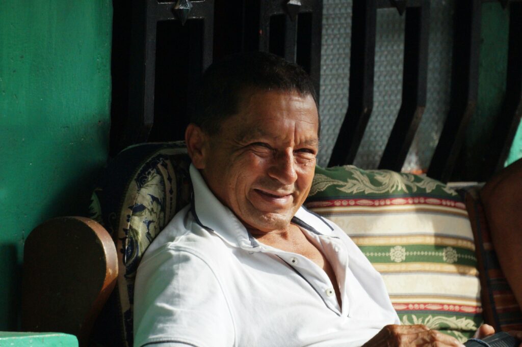 A senior man smiling warmly while sitting outside on a colorful chair.